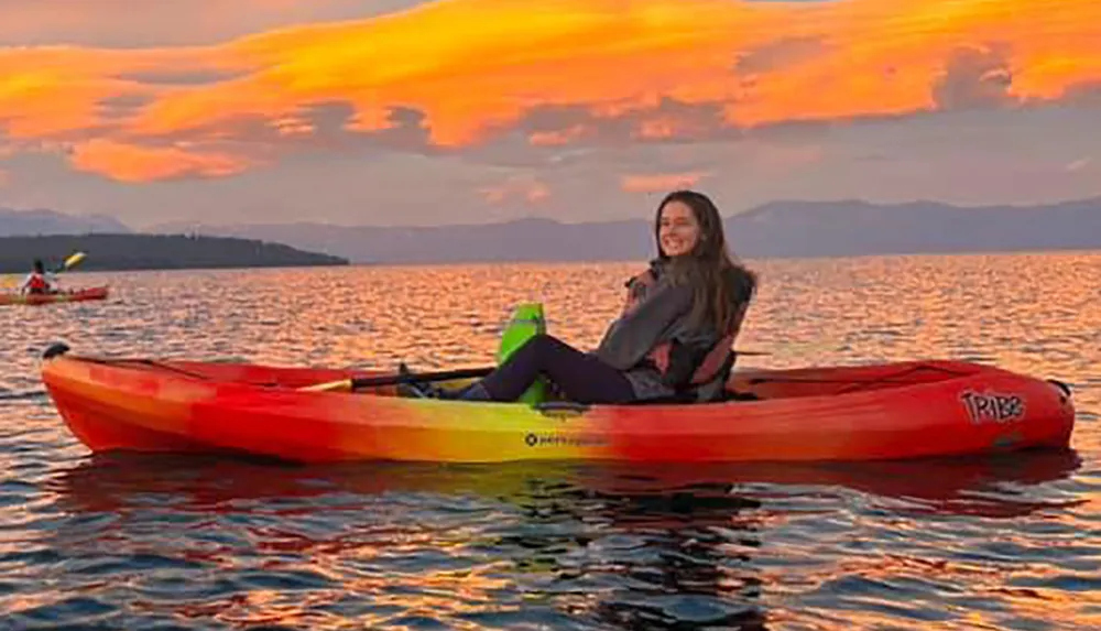 A person smiles while sitting in a vibrant red kayak on calm waters with an orange-hued sunset backdrop