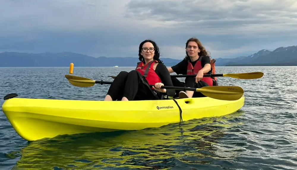 Two individuals are kayaking in a tandem yellow kayak on calm waters with mountains in the distance