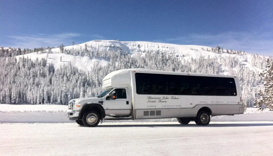 A tour bus is driving along a snowy road with a beautiful backdrop of snow-covered trees and mountains.