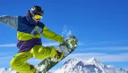 A snowboarder in colorful winter gear is performing a trick on a sunny mountain slope with clear blue skies in the background.