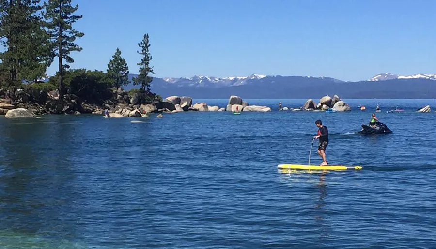 A person is paddleboarding on a clear blue lake with a backdrop of mountains and a jet ski nearby.