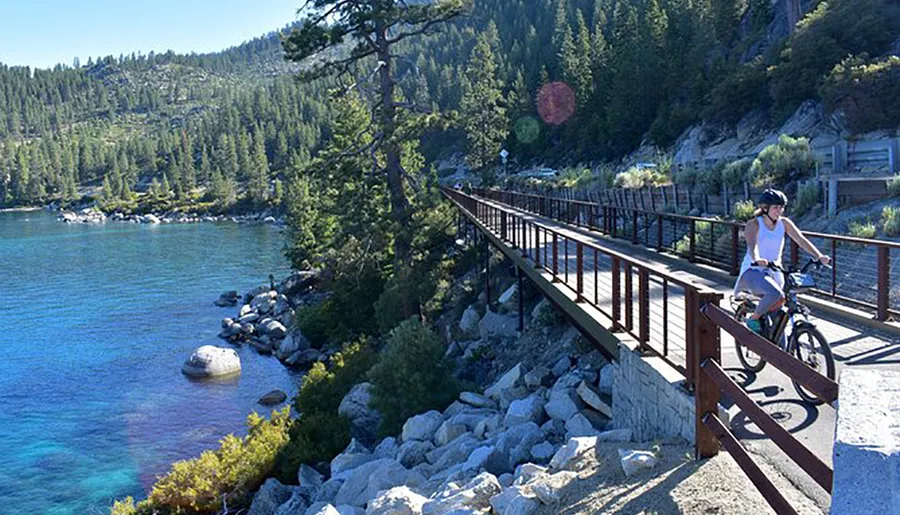 A person is riding a bicycle along a scenic lakeside trail with clear blue water and forested hills in the background.