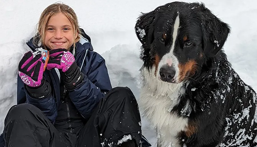 A smiling child in a winter jacket sits in the snow next to a snow-covered Bernese Mountain Dog, both appearing happy and cozy despite the cold surroundings.