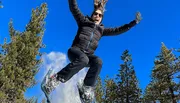 A person is captured mid-jump with a joyful expression, against a clear blue sky backdrop, with snow scattering from their boots.