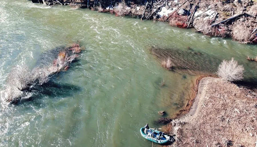 An aerial view of a person in a small boat on a river near a wooded riverbank.