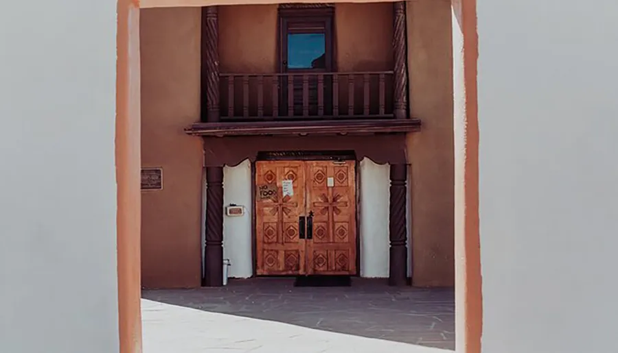 The image shows a traditional Pueblo-style entrance with a carved wooden door and a small upper balcony framed by a terra cotta-colored archway.