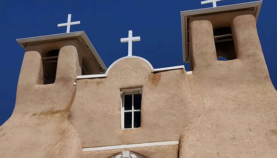 The image shows the upper part of an adobe church with twin bell towers topped with crosses against a blue sky.