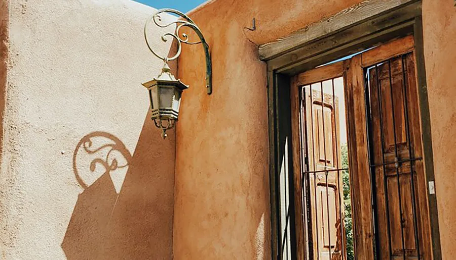 An ornate street lamp casts a decorative shadow on a sunlit, warm-colored adobe wall next to a wooden door.