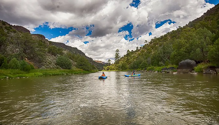 Two people kayak down a serene river flanked by verdant hills and a picturesque cloudy sky.