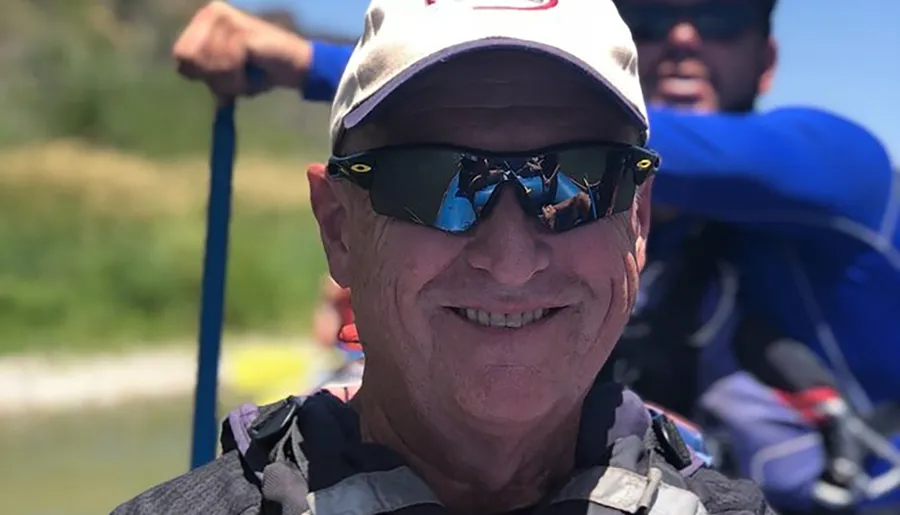 A smiling individual wearing a baseball cap and reflective sunglasses is holding a paddle, suggesting they are engaged in an outdoor water activity, with another person visible in the background.