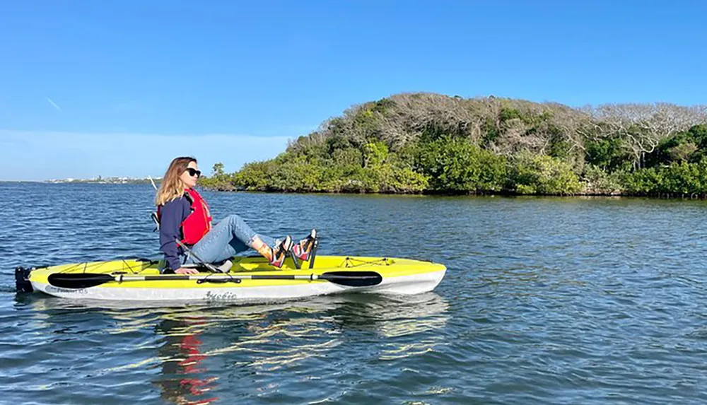 A person is casually seated on a yellow kayak enjoying the calm waters near a green-covered coastline under a clear blue sky