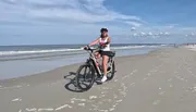 A person is standing with a bicycle on a sandy beach on a sunny day, with the ocean in the background.