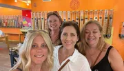 Four smiling women take a selfie together in a colorful candy store with dispensers filled with various sweets in the background.