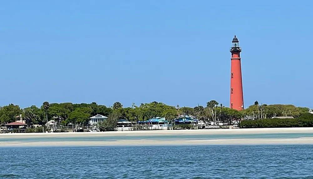 A tall red lighthouse stands strikingly beside waterfront buildings against a clear blue sky