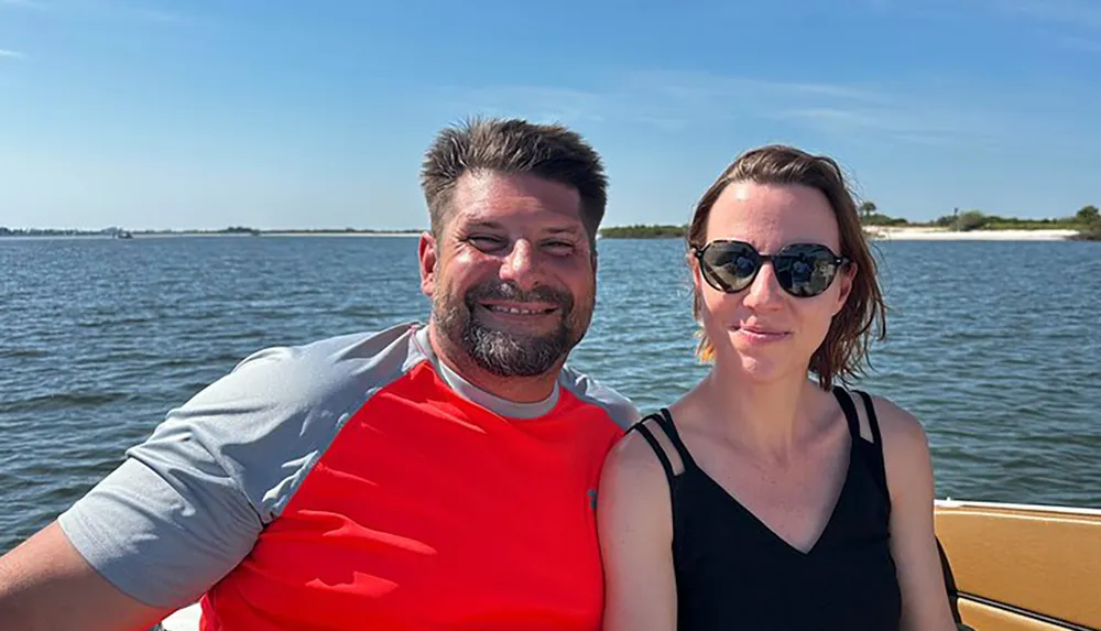 Two people are smiling for a photo while enjoying a sunny day on a boat with calm waters and a clear sky in the background