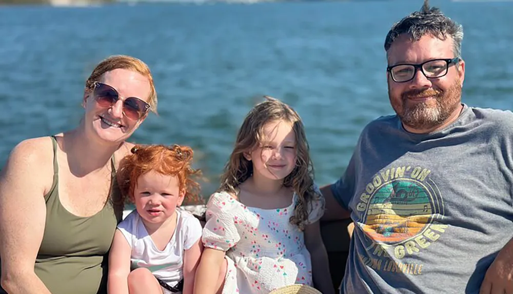 A family of four is smiling and posing together by the water on a sunny day