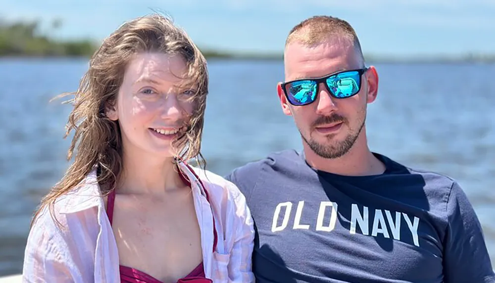 A man and a woman are smiling for a photo with a body of water in the background on a sunny day