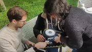 Two people are attentively examining an electronic device outdoors.