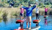 Four people with paddles raised high are standing on paddleboards in a waterway surrounded by grass, with one person having a small dog on their board.