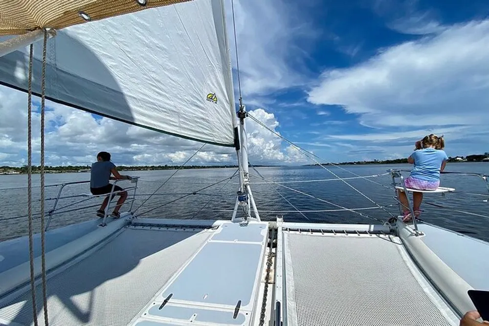 Two individuals are enjoying a peaceful day sailing on a catamaran under a partly cloudy sky