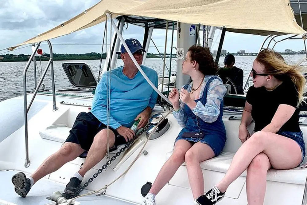 Three individuals are engaging in a conversation on a boat with a body of water and overcast skies in the background