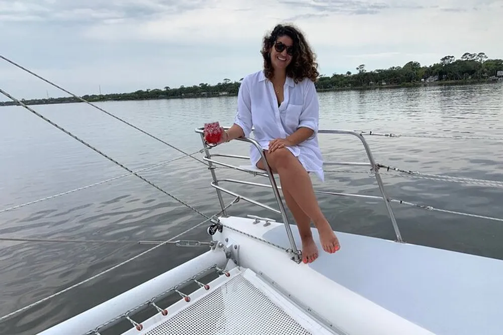 A smiling person is sitting relaxed on the bow of a sailboat holding a drink with calm waters and greenery in the background