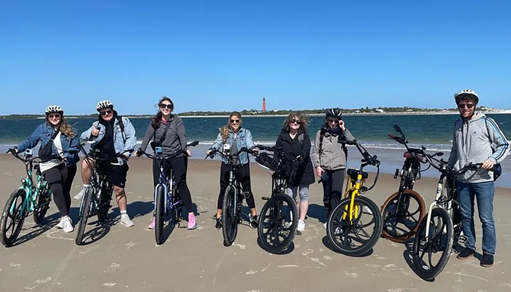 A group of seven people wearing helmets pose with their bicycles on a sandy beach with a blue sky above and a lighthouse visible in the distance