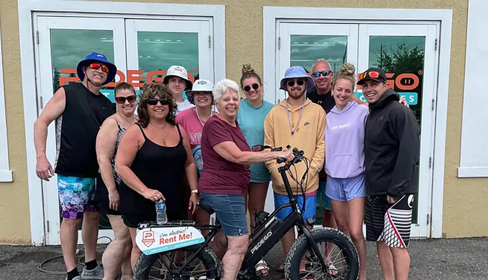 A group of people of varying ages are smiling for a photo in front of a bike rental shop with some wearing summer attire and accessories suggesting they are on a holiday or enjoying a recreational outing