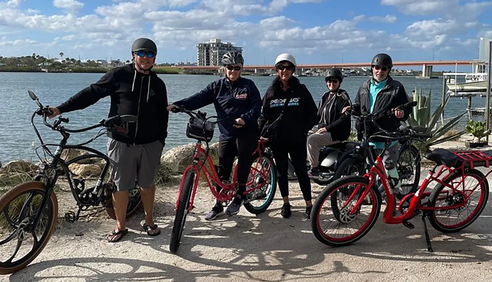 A group of five people each with a bicycle are posing for a photo by a waterfront with a bridge in the background