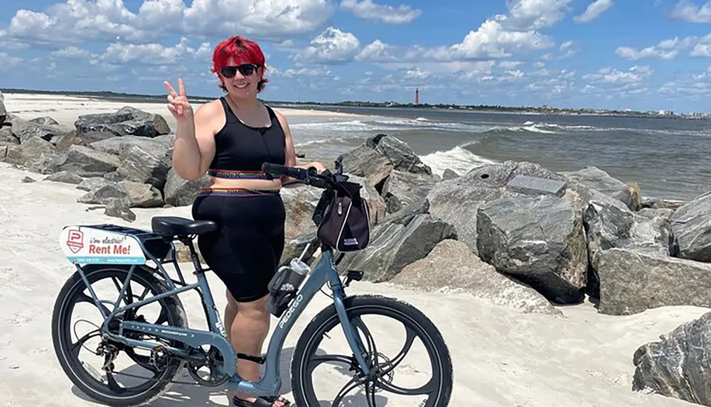 A person poses with a peace sign next to a rental bike on a sandy beach with rocks and a distant lighthouse in the background