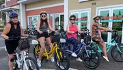 Four people are smiling and posing with colorful electric bikes in front of a bike rental shop.