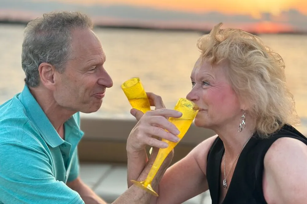 A couple is playfully toasting with yellow plastic cups against a backdrop of a serene sunset over the water