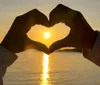 Two hands form a heart shape against a sunset over water creating a silhouette with the sun in the center