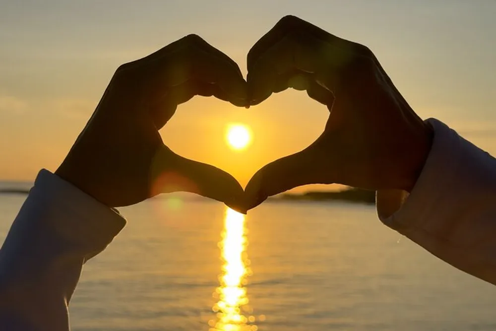 Two hands form a heart shape against a sunset over water creating a silhouette with the sun in the center