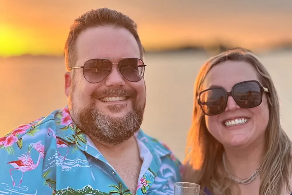 Two smiling individuals in sunglasses are enjoying a beautiful sunset by the water