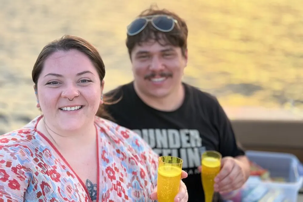 A smiling woman and a man with sunglasses atop his head are enjoying drinks outdoors with a blurred natural backdrop suggesting a relaxed convivial setting