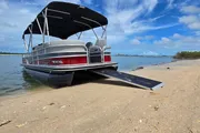 A pontoon boat is docked on a sandy shore under a blue sky with scattered clouds.