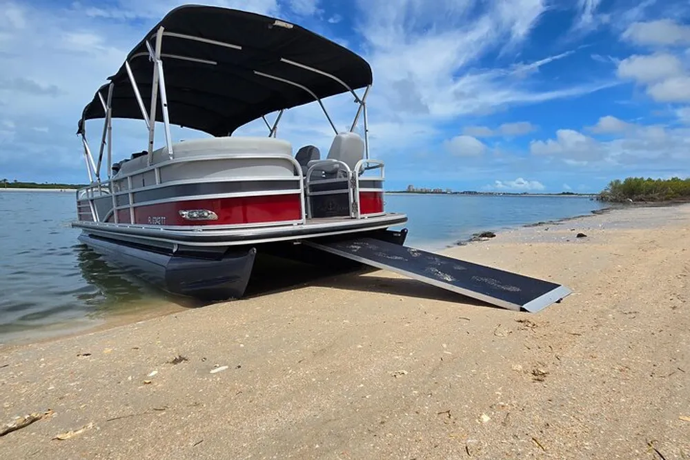 A pontoon boat is docked on a sandy shore under a blue sky with scattered clouds