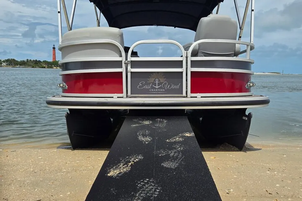 A pontoon boat is docked on a sandy shore with a lighthouse visible in the background