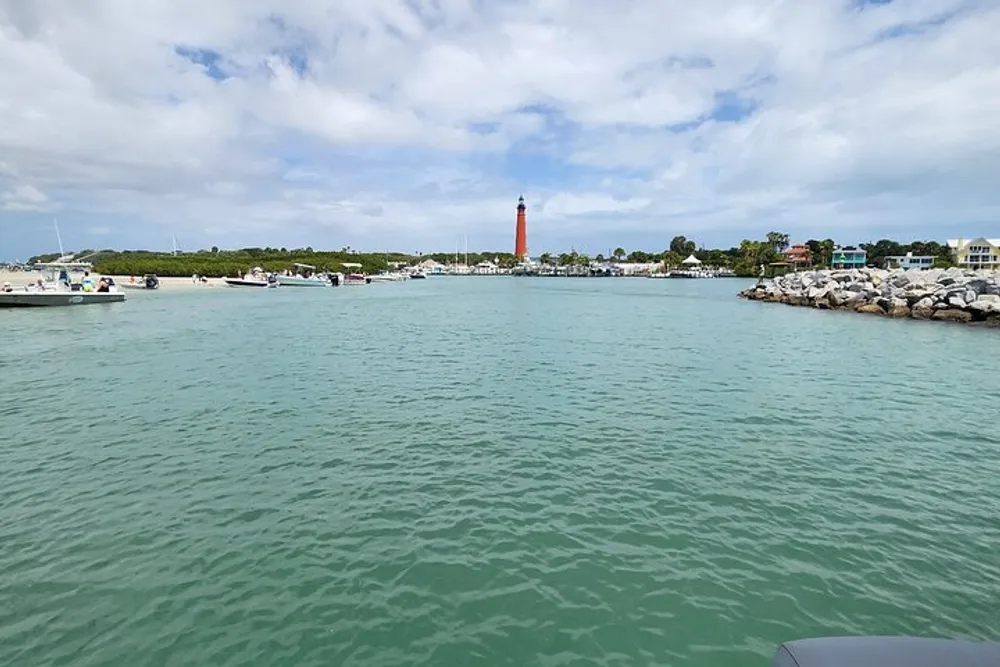 The image shows a coastal scene with a red lighthouse in the background boats moored in calm turquoise waters and a rocky jetty on the right under a partly cloudy sky