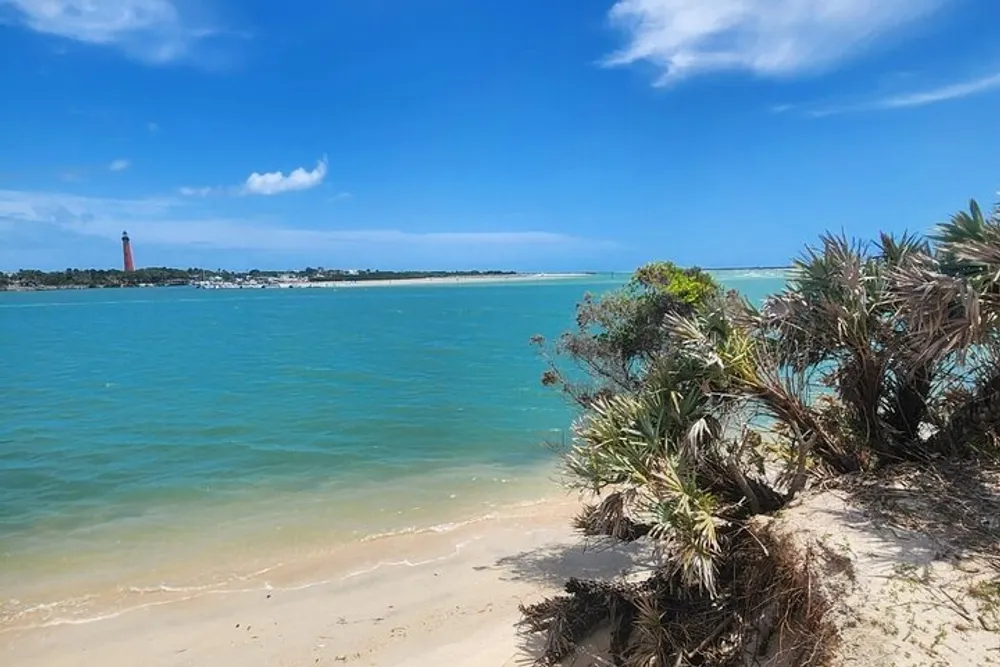 The image shows a scenic beach view with clear turquoise waters white sand lush green vegetation in the foreground and a tall red and white lighthouse in the distance under a blue sky with fluffy clouds