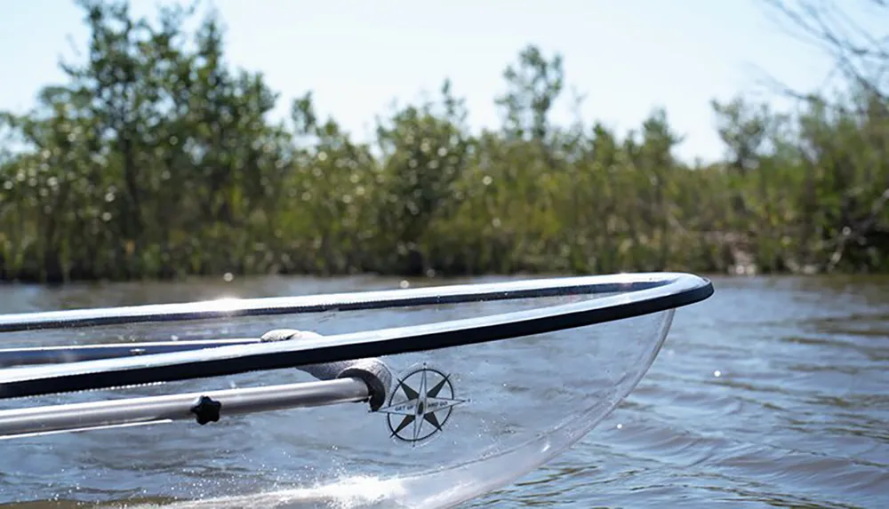 This image shows the bow of a clear kayak gliding through water with greenery in the background highlighting a unique perspective on kayaking