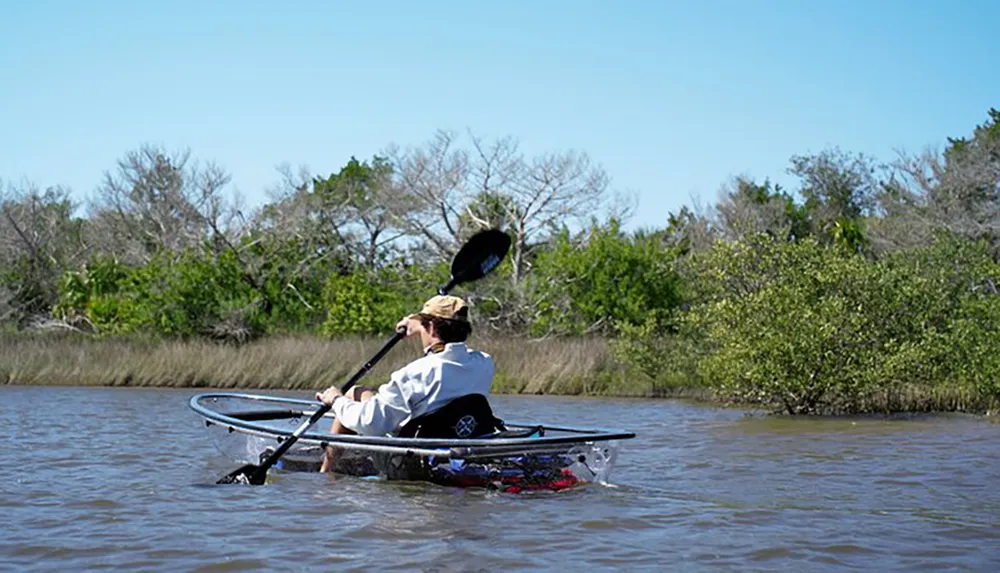 A person wearing a hat paddles a kayak through calm waters surrounded by greenery