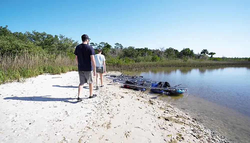 Two individuals are walking alongside a tranquil body of water where kayaks are rested on the sandy shore under a clear blue sky