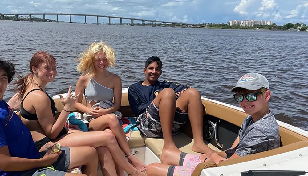 A group of young people are enjoying a boat ride on a sunny day with a bridge and buildings in the background