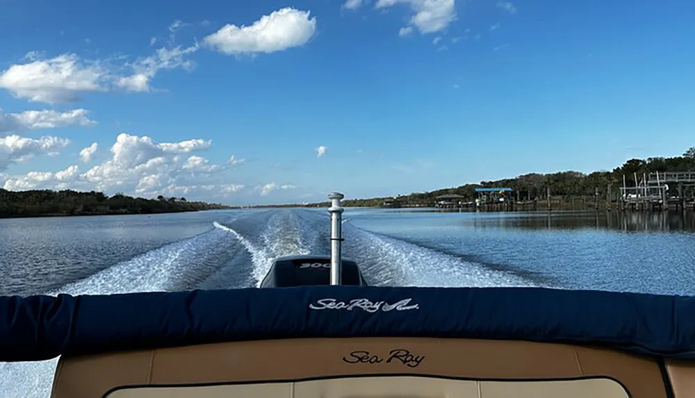 A boat named Sea Ray is cruising on a calm river leaving a wake behind under a partly cloudy sky