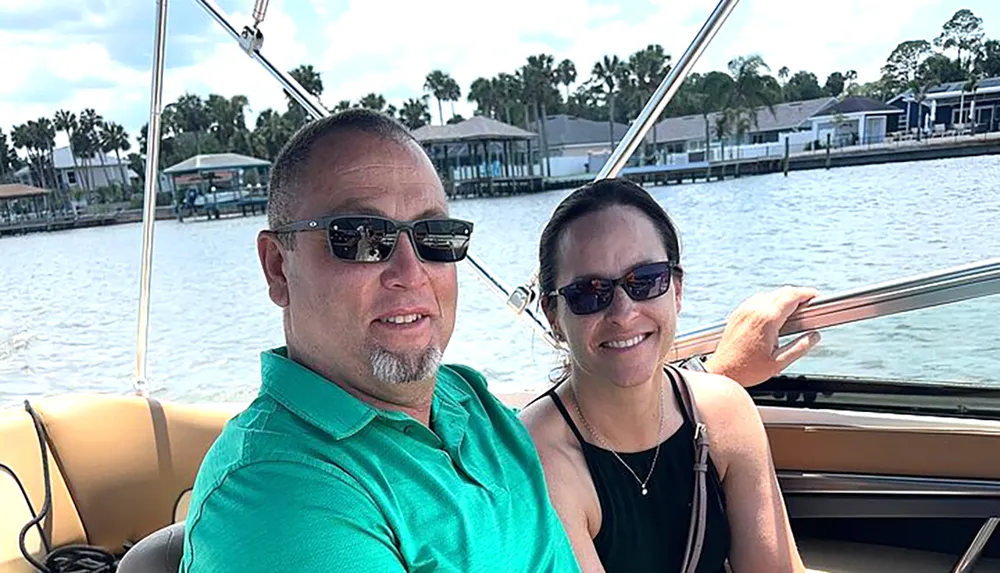 Two people are smiling for a selfie on a boat with waterfront properties in the background