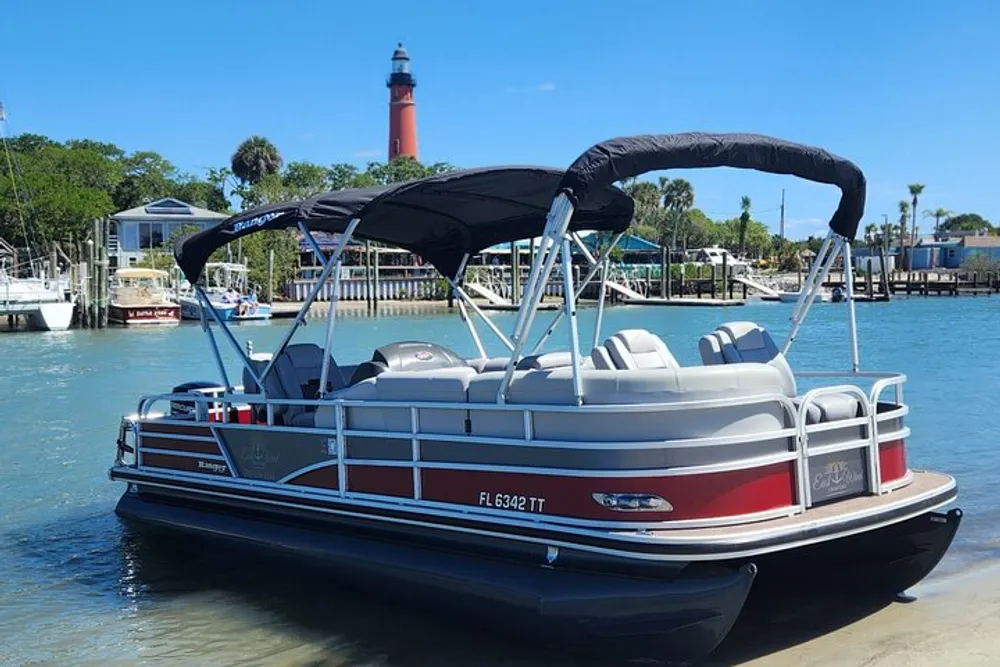 A pontoon boat is anchored in calm waters near a dock with a tall red lighthouse in the background under a clear blue sky