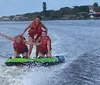 Three people are wearing life jackets excitedly riding a towable water tube behind a boat with coastal buildings in the background