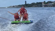 Three people are wearing life jackets, excitedly riding a towable water tube behind a boat, with coastal buildings in the background.